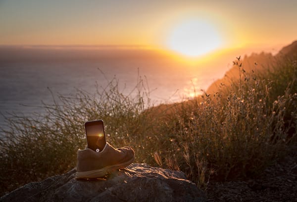 Schuh mit Mobiltelefon auf einem Felsen vor einer Kuestenlandschaft im Sonnenuntergang