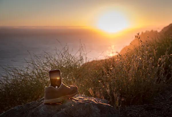 Schuh mit Mobiltelefon auf einem Felsen vor einer Kuestenlandschaft im Sonnenuntergang