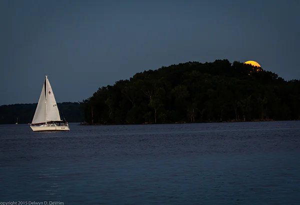 Segelboot auf dunklem Wasser vor einer Insel mit aufgehendem Mond